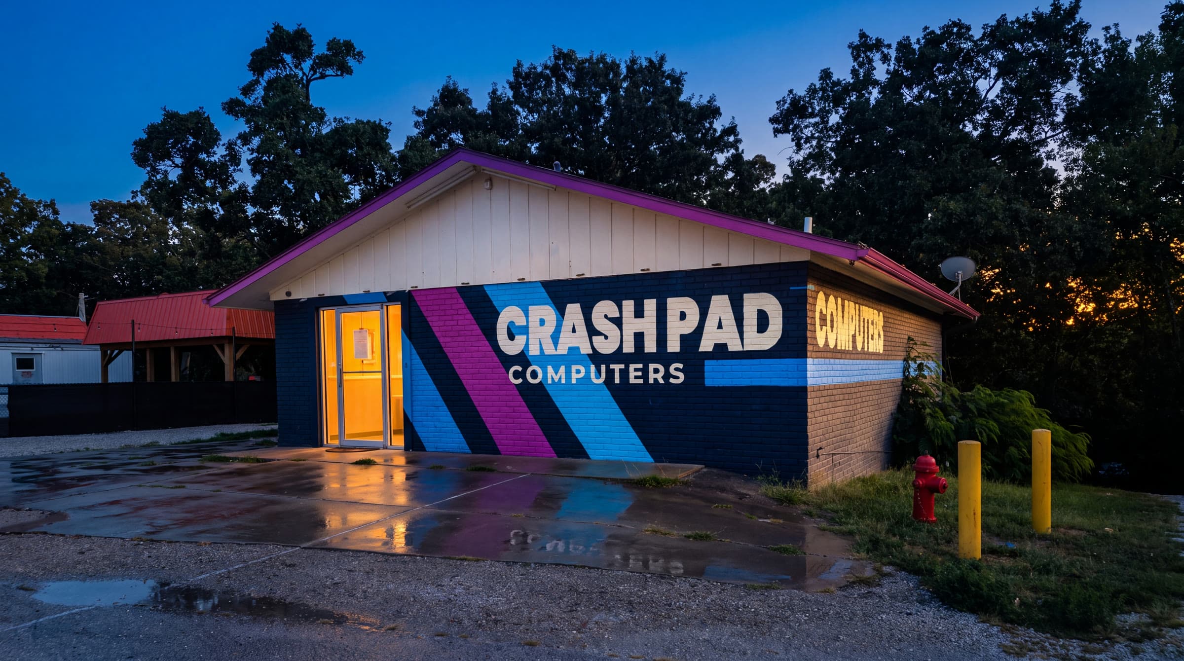 The Crash Pad Computers building on Route 5 — navy brick with the painted stripe mural. Photographed at dusk from the lot on 14327 MO-5.
